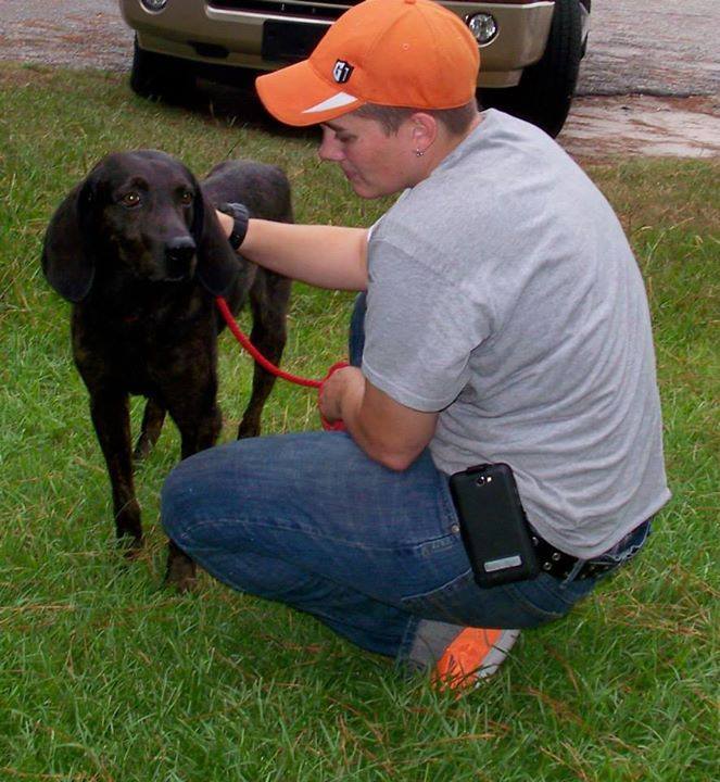Trainer kneeling in the grass offering calm reassurance to a nervous hound on leash, reflecting relationship-based dog training.